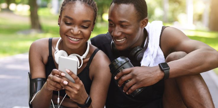 a happy couple in athletic wear enjoy a break, smiling and looking at a phone together outdoors.