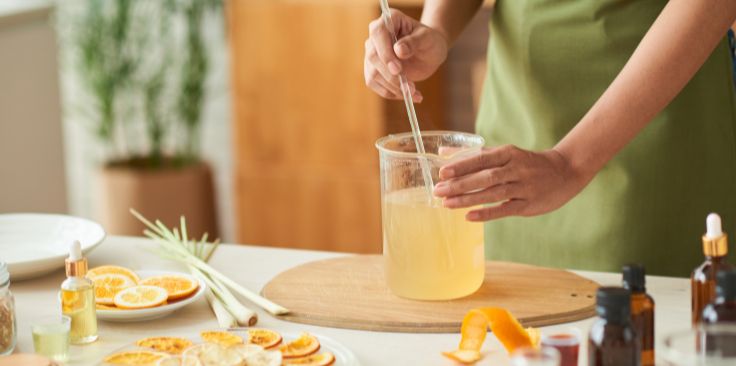 a person stirring a glycerin mixture in a beaker, surrounded by citrus slices and essential oils.