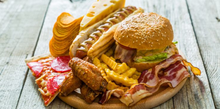 a plate of unhealthy high-fat foods including a cheeseburger, hotdog, bacon, fried chicken wings, French fries, pizza, and potato chips on a wooden surface.