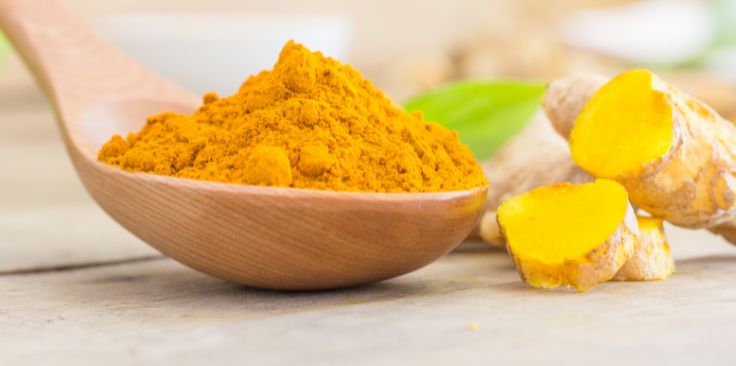 a wooden spoon filled with bright yellow turmeric powder next to fresh turmeric roots.