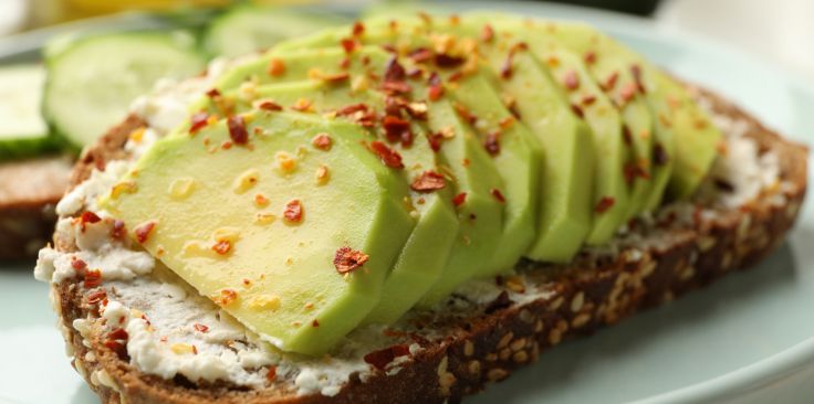 multigrain toast topped with creamy avocado slices, red pepper flakes, and a white spread, with cucumber slices in the background.