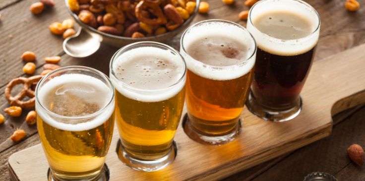 a flight of four beers on a wooden board with a bowl of mixed nuts in the background.