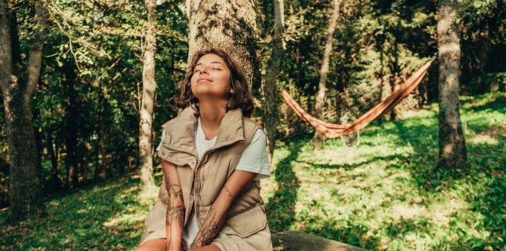 a woman peacefully enjoying nature, sitting against a tree with a hammock in the background.