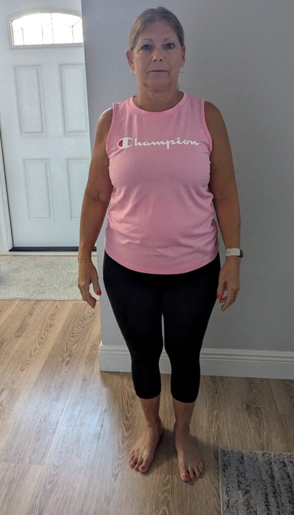 a woman standing indoors in a pink tank top and black leggings, with her hair tied back, looking straight at the camera.