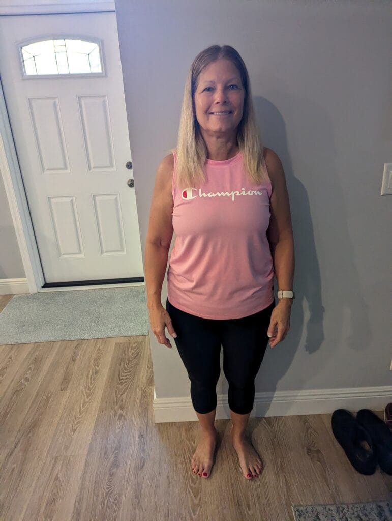 A woman standing indoors in a pink tank top and black leggings, with her hair down, smiling and facing the camera.