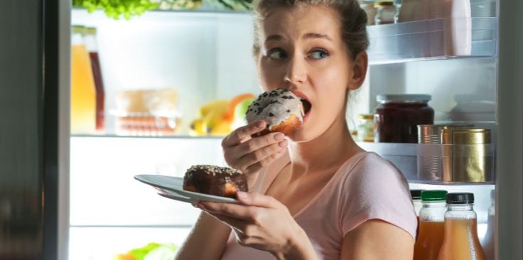 a woman eating a donut while standing in front of an open refrigerator.