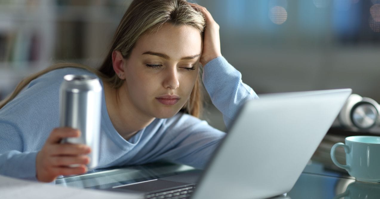 Are Energy Drinks Bad for Your Liver: Fatigued woman holding an energy drink in front of a laptop.