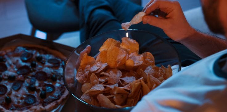 Person eating chips and pizza while sitting on a couch at night.