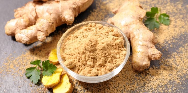 fresh ginger root with a bowl of ground ginger powder on a slate background.