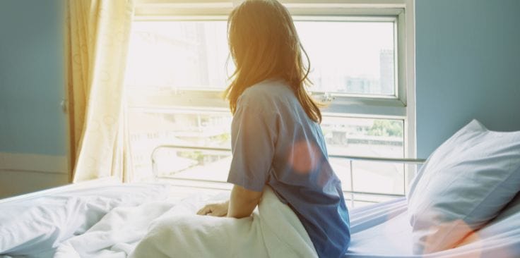 A woman sitting in a hospital bed by the window with sunlight streaming in.