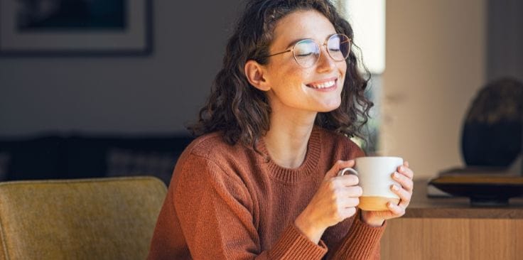 Smiling woman wearing glasses enjoying a warm cup of tea at home.