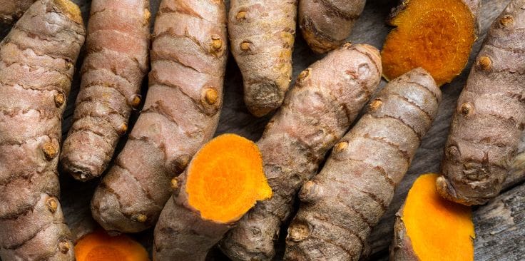 a close-up view of fresh turmeric roots, showcasing their earthy brown outer skin and vibrant orange interior, arranged on a rustic wooden surface.