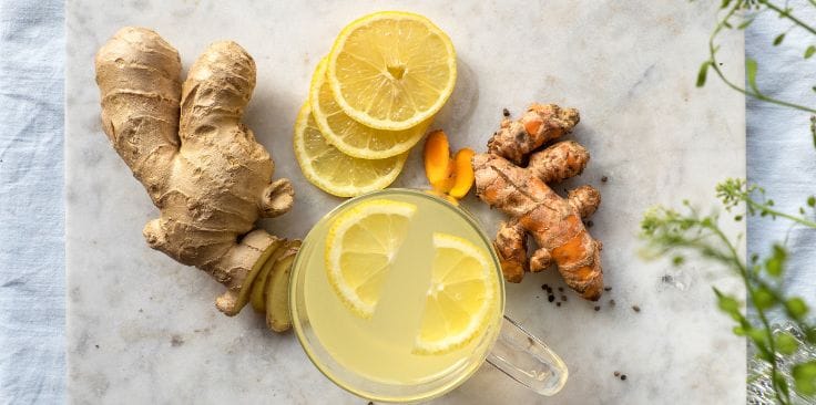 Fresh ginger, turmeric, lemon slices, and a cup of turmeric ginger tea on a marble surface.