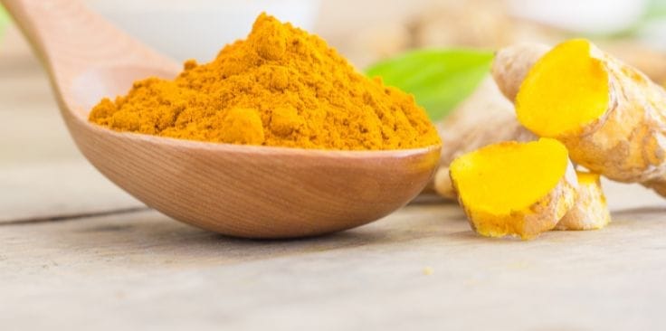 vibrant yellow turmeric powder in a wooden spoon, accompanied by fresh turmeric roots sliced to reveal their rich orange interior, placed on a wooden surface with green leaves in the background. 