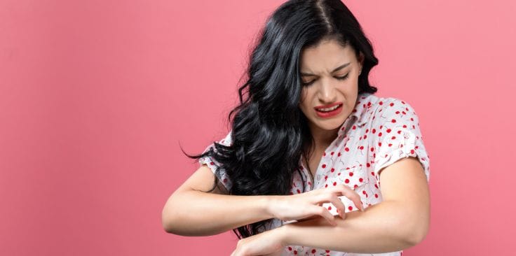 woman scratching an itchy rash on her arm with a frustrated expression.