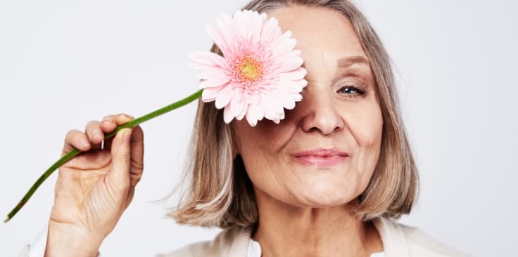 a confident older woman playfully holds a pink daisy to her eye, radiating positivity and joy.