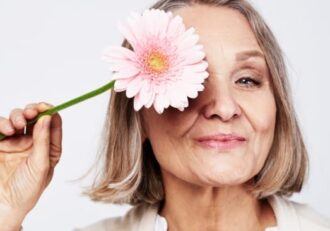 a confident older woman playfully holds a pink daisy to her eye, radiating positivity and joy.
