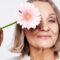 a confident older woman playfully holds a pink daisy to her eye, radiating positivity and joy.